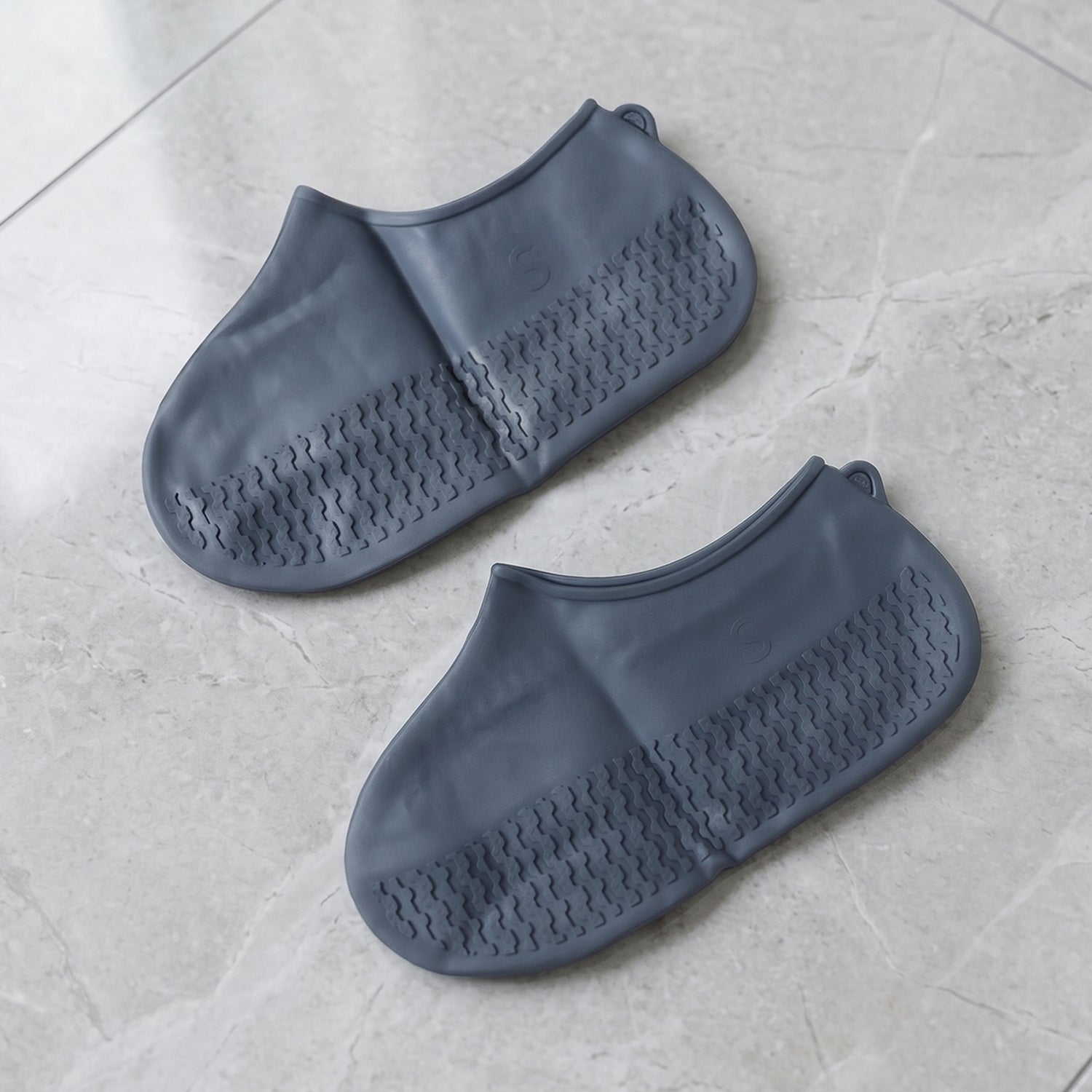 A pair of dark gray silicone shoe covers with textured soles is presented on a light gray marble-patterned floor.